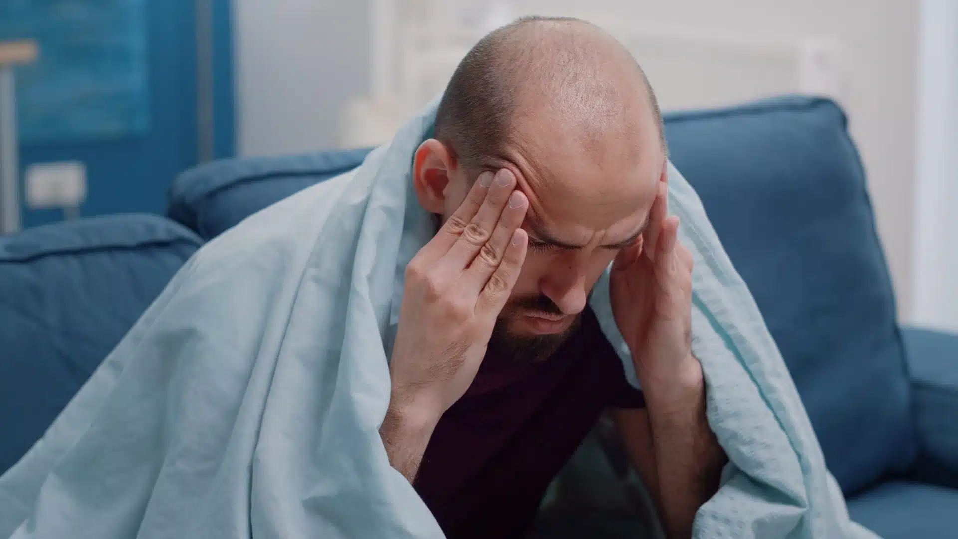 Man with headache and fatigue sitting on couch covered with blanket pressing temples