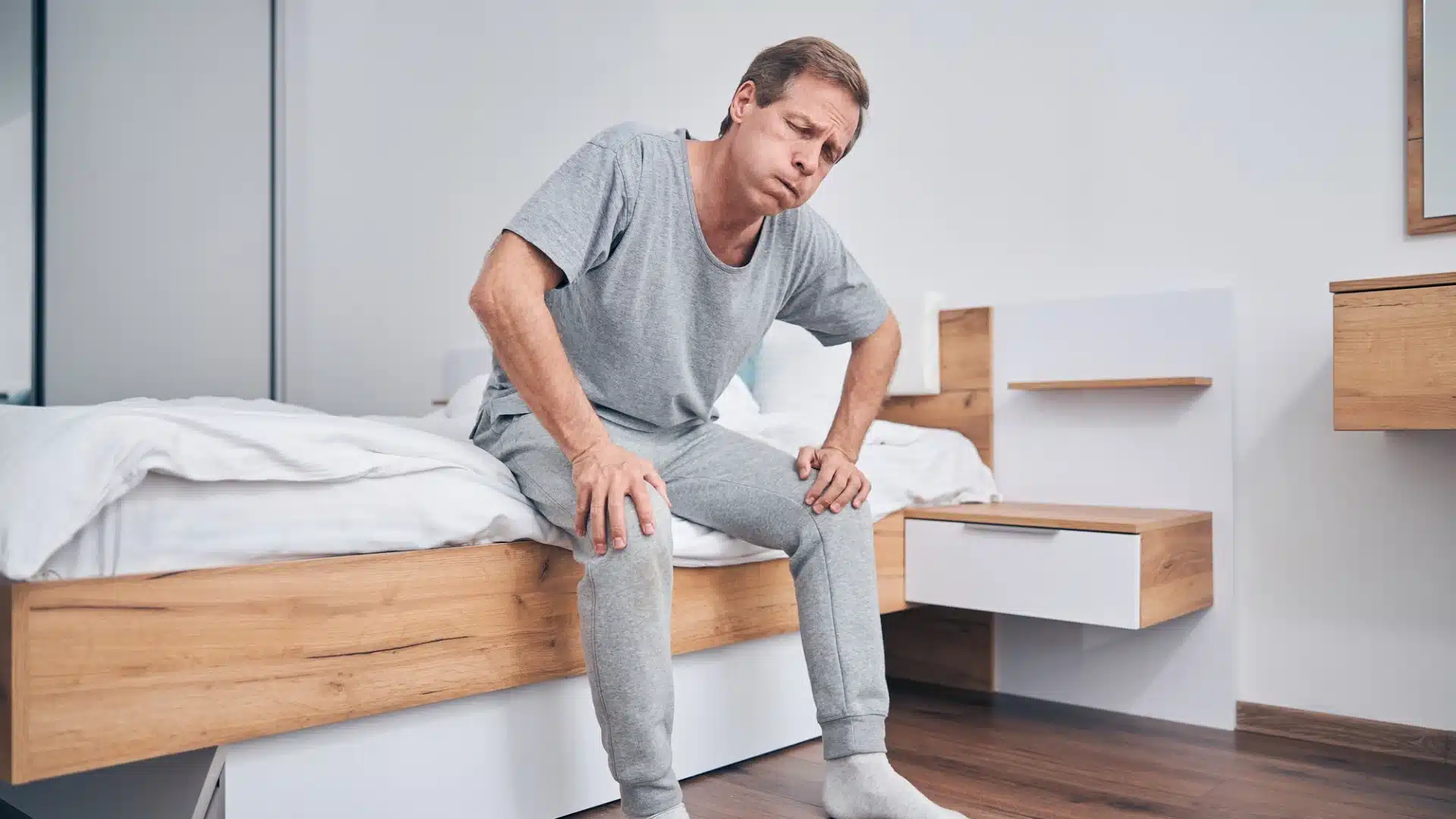 Middle-aged man sitting on bed holding knees, suffering from joint pain and stiffness in the morning
