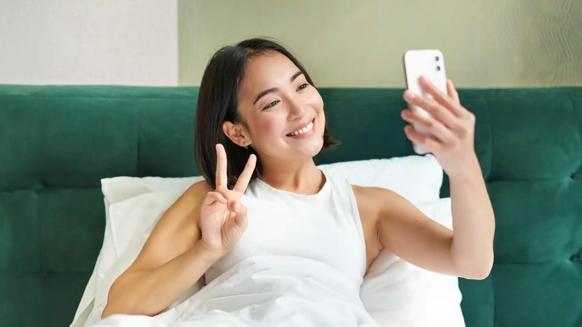 Smiling woman in bed taking selfie with peace sign