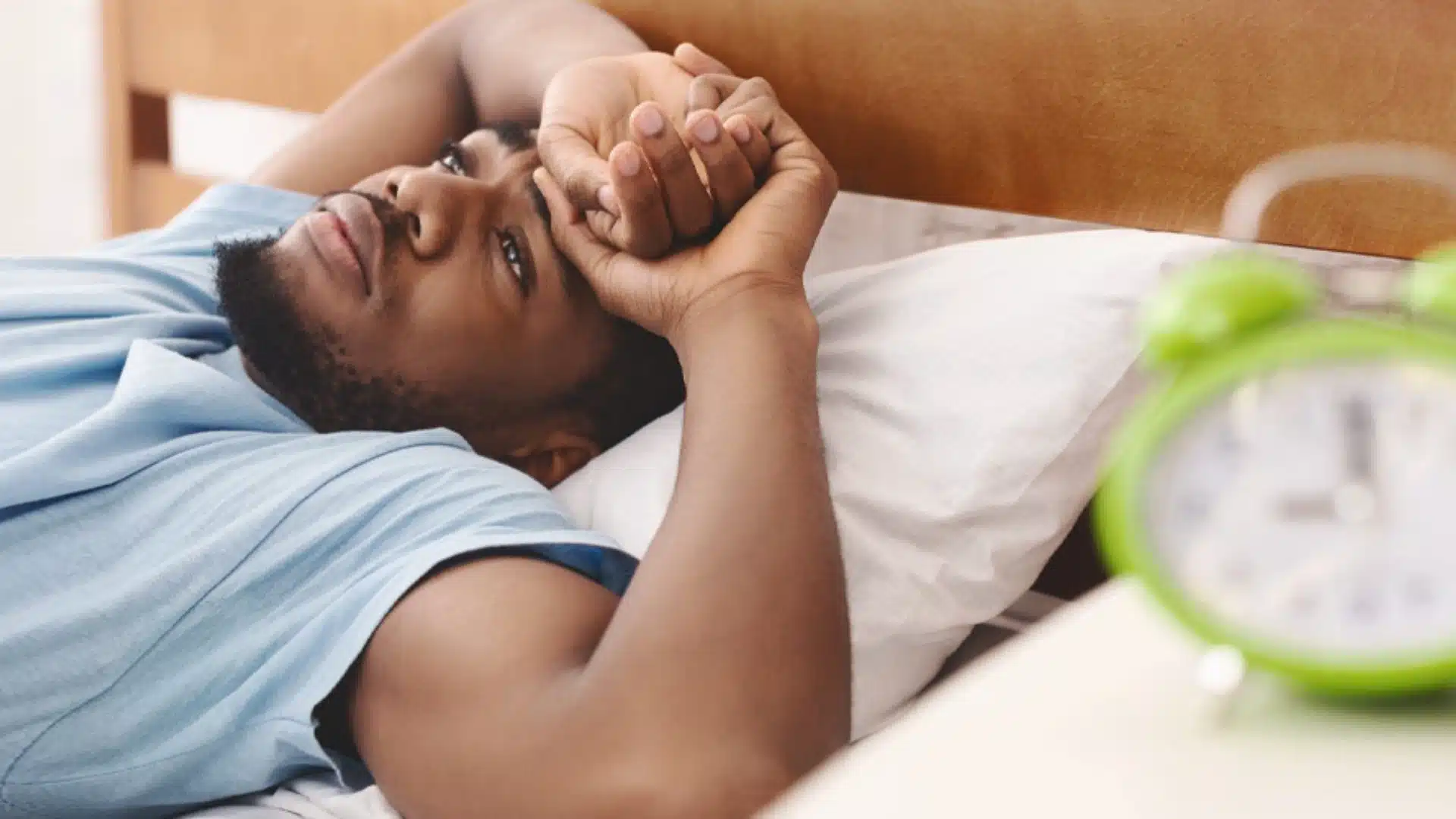 Young man lying awake in bed with alarm clock in foreground, experiencing insomnia or sleep disturbance