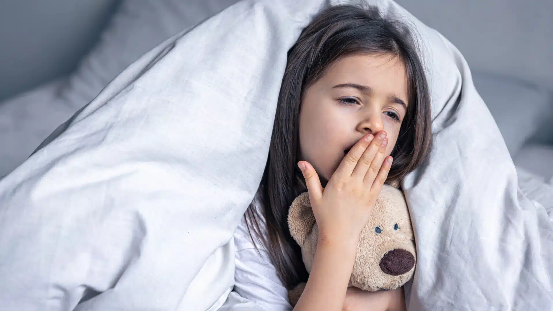 Sleepy little girl yawning in bed while holding teddy bear under white blanket