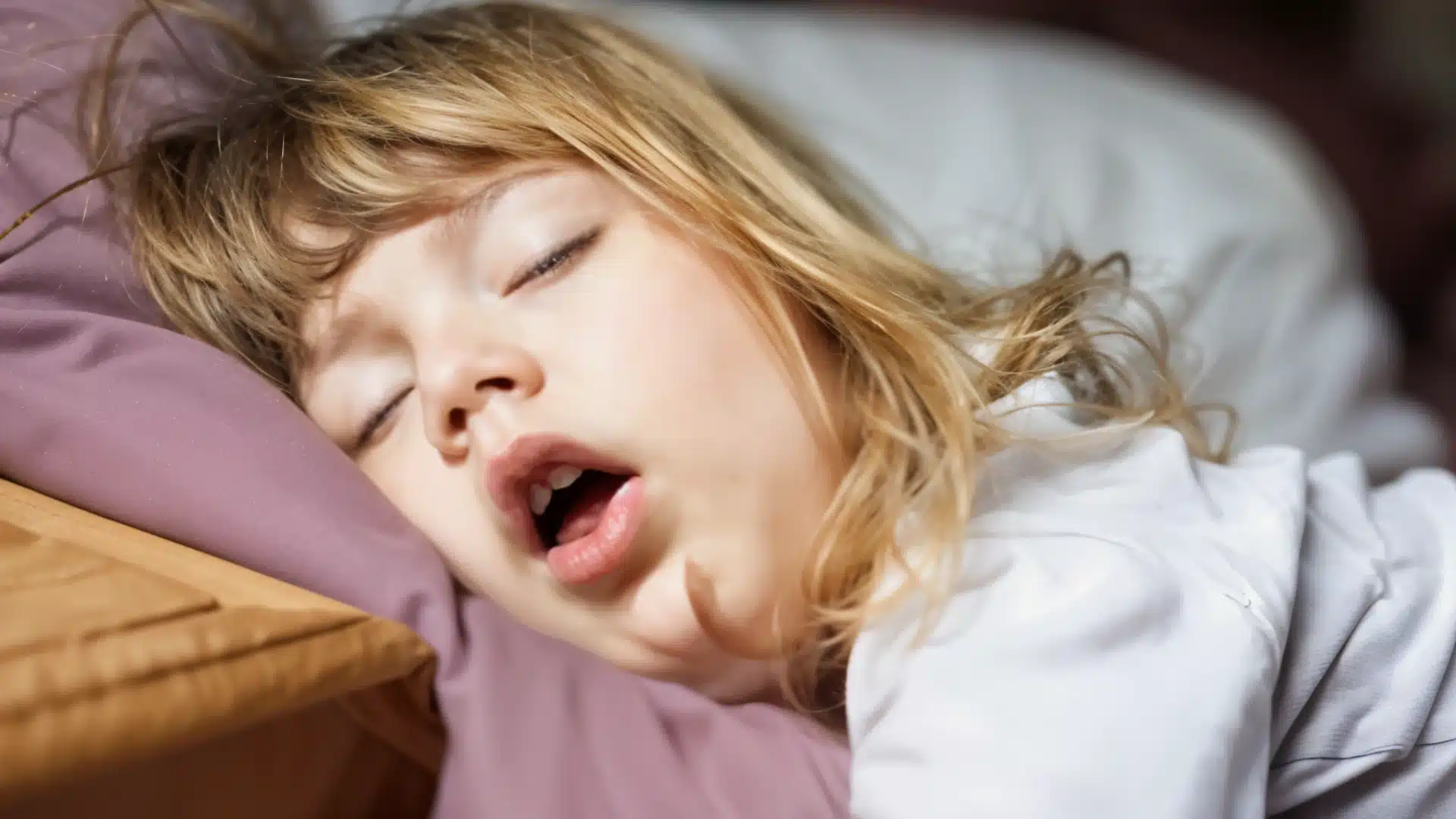 Young child sleeping with mouth open on pillow, showing signs of possible snoring or sleep apnea in kids