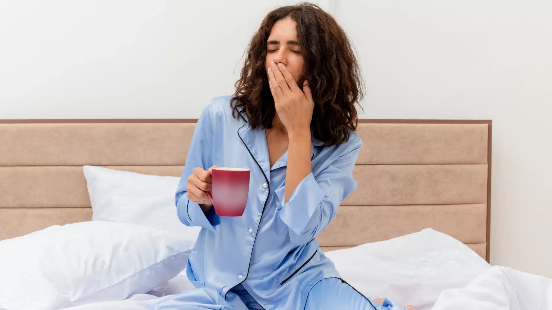 Tired woman in blue pajamas yawning and holding coffee cup in bed