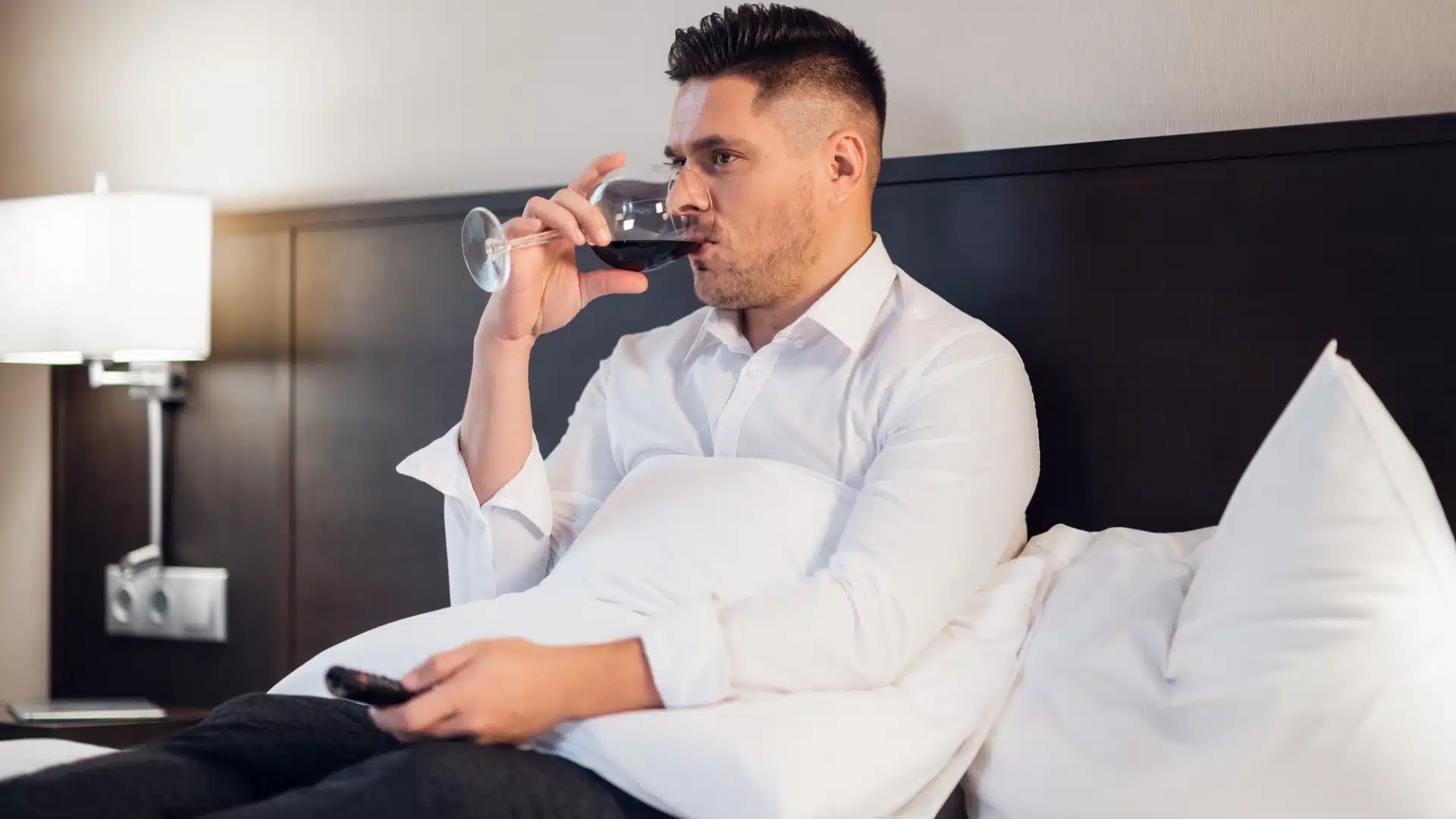 Man sitting in bed drinking red wine while watching TV in the evening