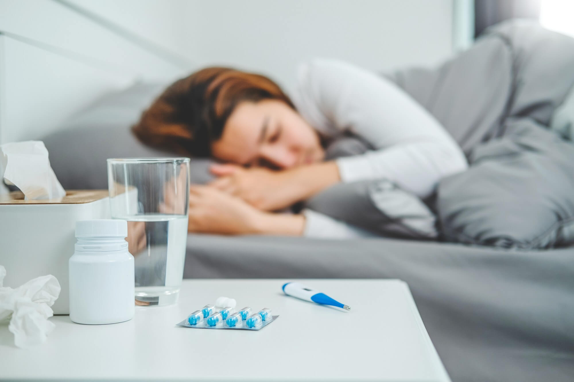 Sick woman resting in bed with medicine, glass of water, and thermometer on bedside table