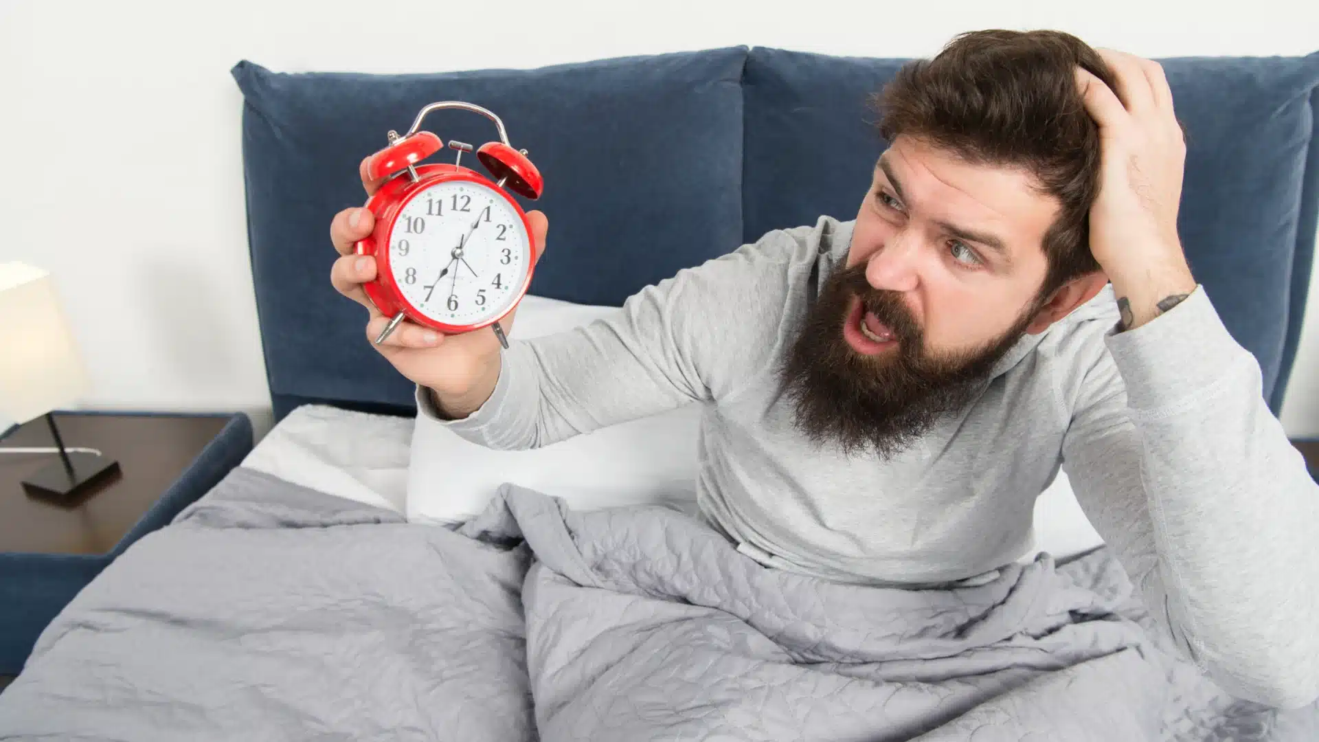 Shocked man waking up late in bed holding red alarm clock and looking stressed