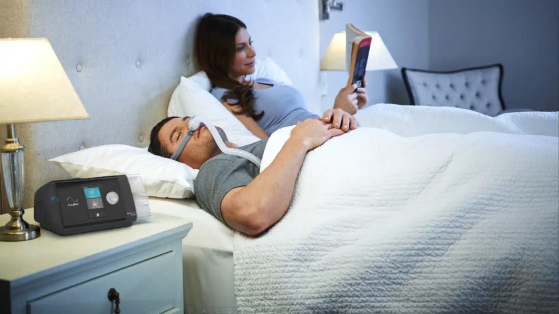 Man sleeping with CPAP machine for sleep apnea treatment while partner reads in bed