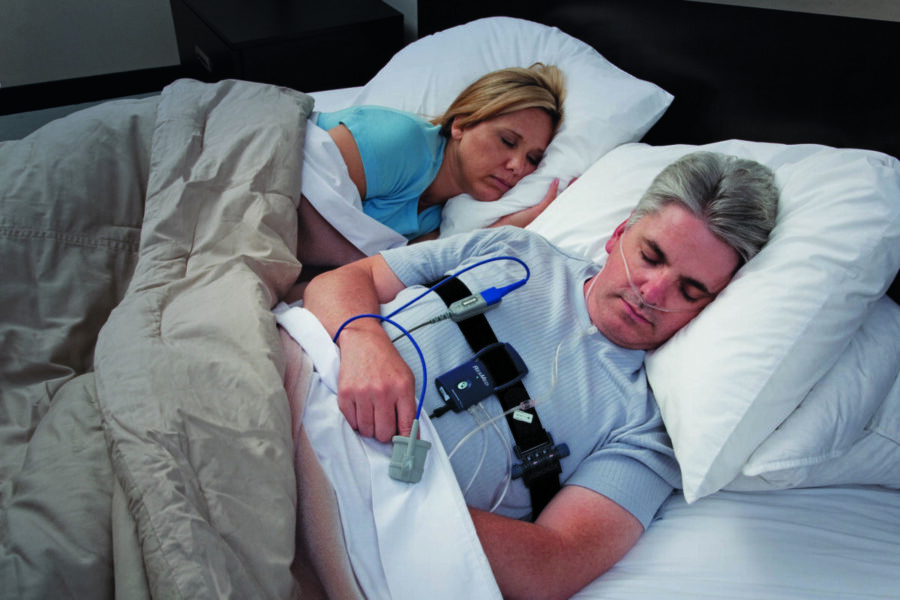 Man undergoing sleep apnea test with monitoring equipment while sleeping beside partner