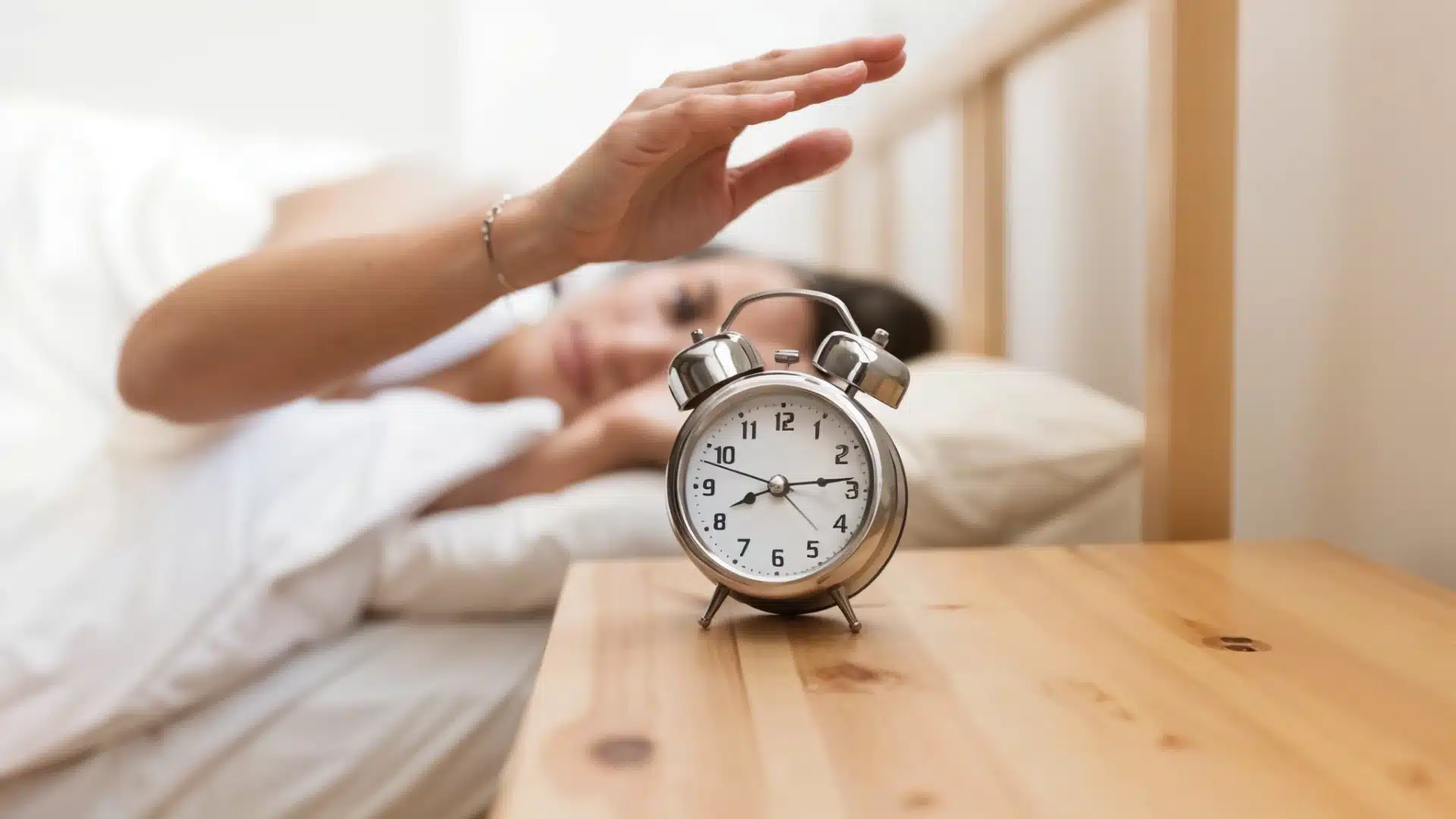 Person in bed reaching to turn off alarm clock on wooden nightstand in the morning